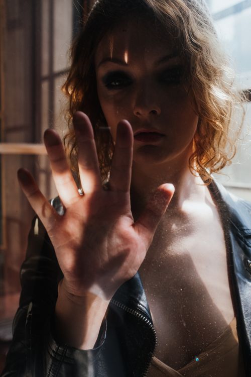 Free A stylish portrait of a woman with smoky eyes behind a window, wearing a leather jacket, indoors. Stock Photo