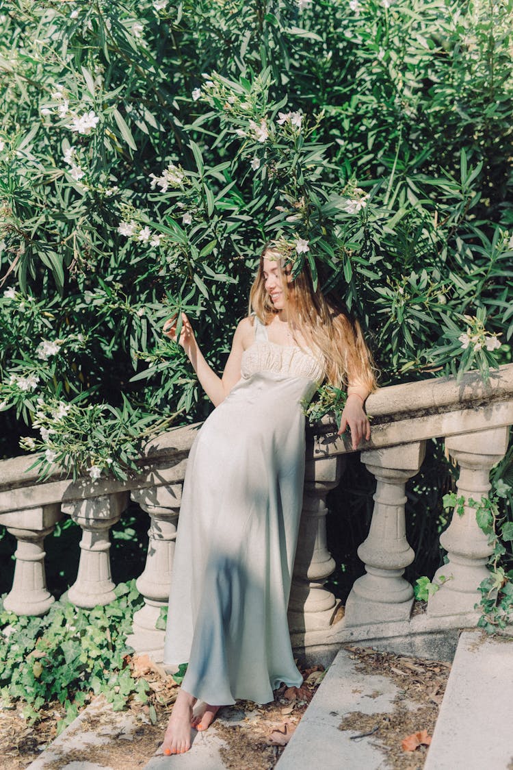 A Woman In White Dress Leaning On Banister Near A Flowering Tree