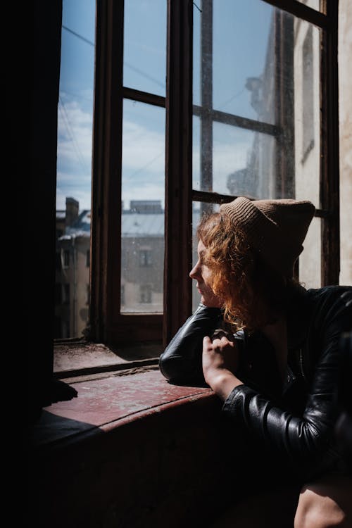 Free Woman in Black Leather Jacket and Brown Knit Cap Sitting Beside Window Stock Photo