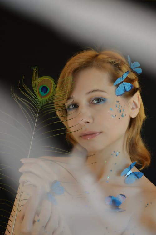 Dreamy female with bare shoulders and blue butterflies on head and body looking at camera while standing in studio with light stain