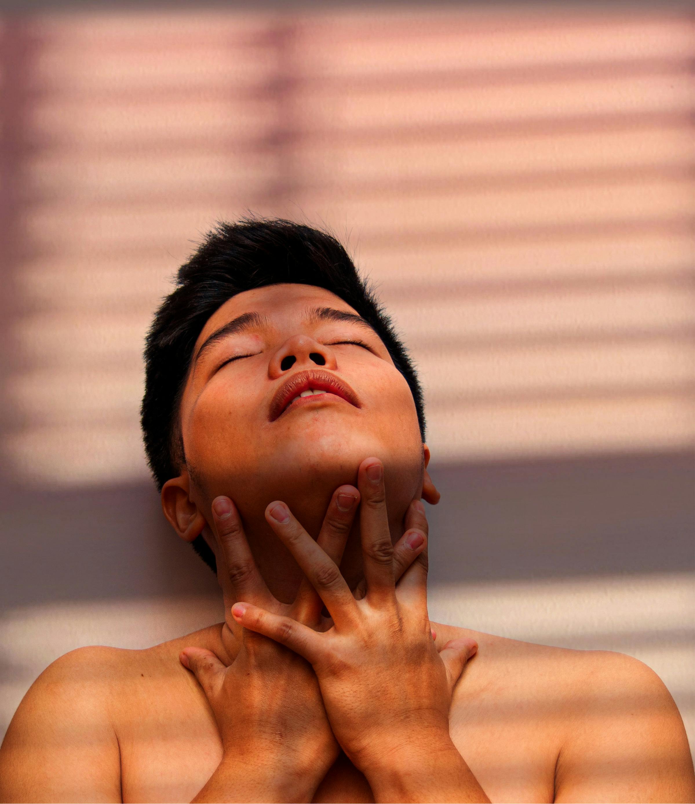 Sensitive Asian male with closed eyes and naked torso touching face while standing near white wall with shadow and sunshine