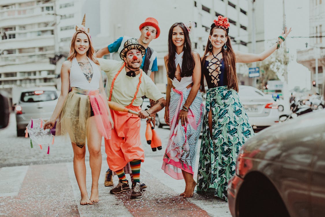 Free Male clowns in bright apparel and hats standing near cheerful girlfriends in ornamental clothes on pavement while looking at ไอเดียจัดกิจกรรมปีใหม่ camera Stock Photo