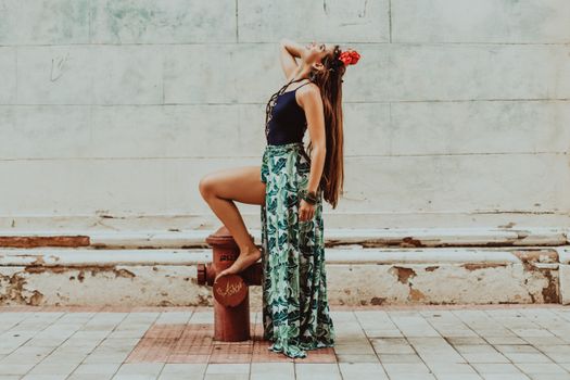 Side view of young content barefoot female in ornamental clothes and floral adornment on head looking up while touching hair on street