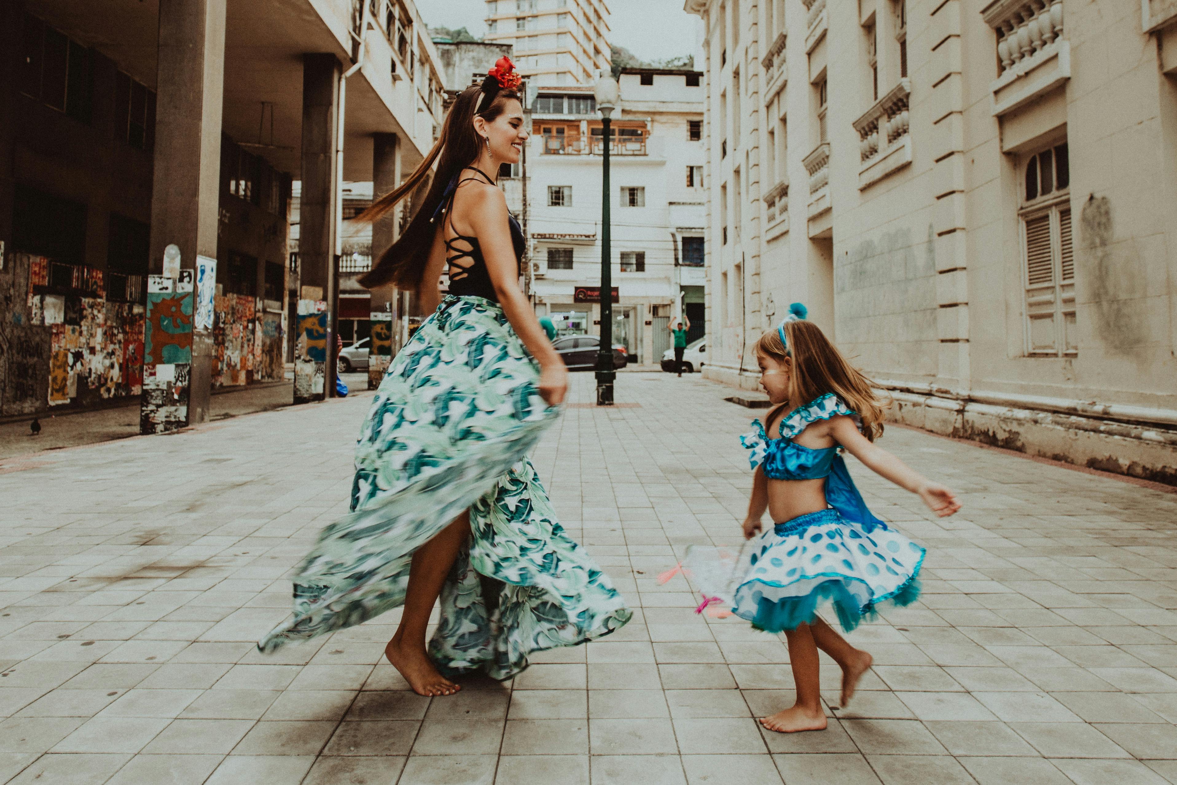 A mother and daughter enjoying a playful, carefree dance in an urban street wearing colorful dresses.