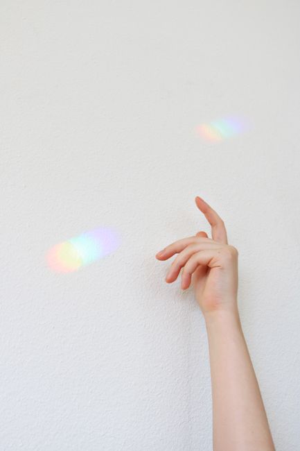 Close-up of a hand reaching toward rainbow reflections on a white wall. Minimalist and artistic.