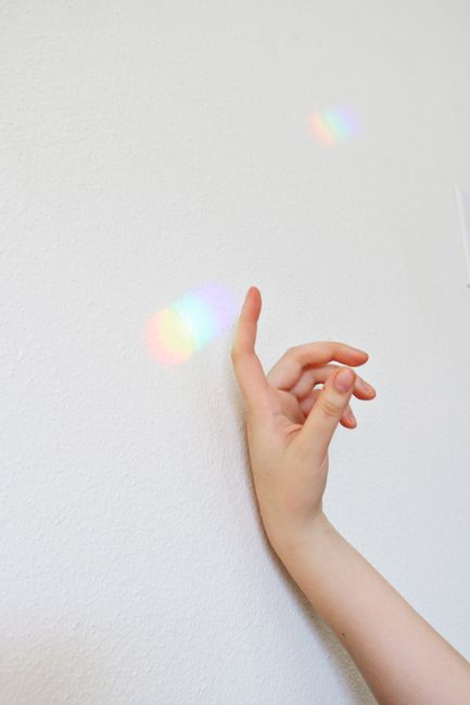 A delicate hand reaches out to touch a vibrant rainbow reflection on a blank white wall.