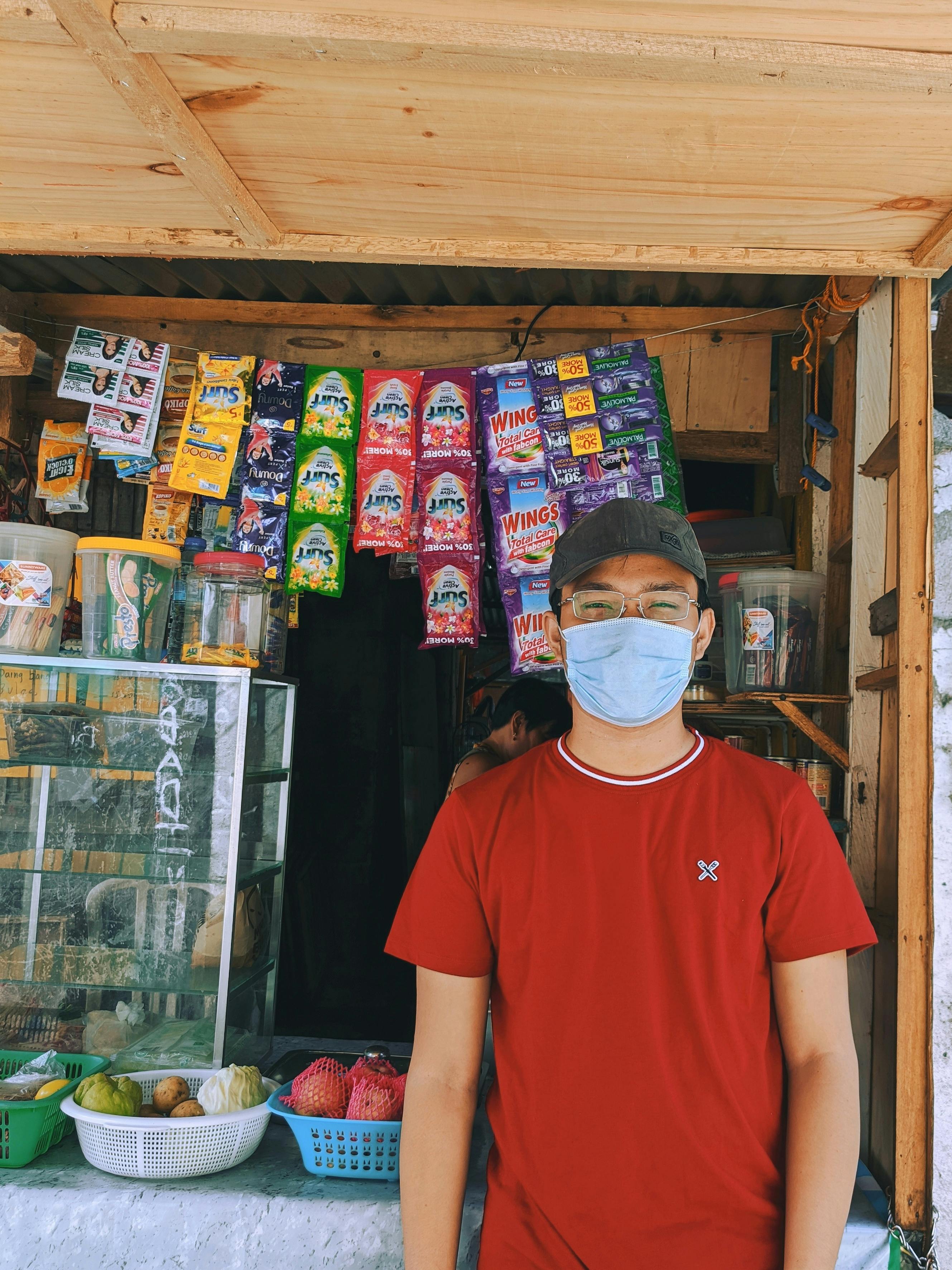 Man wearing mask and glasses standing in front of a small store with diverse goods for sale.