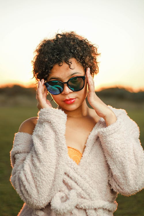 Young confident African American female with short curly hair in soft bathrobe adjusting sunglasses while standing on grassy lawn at sunset
