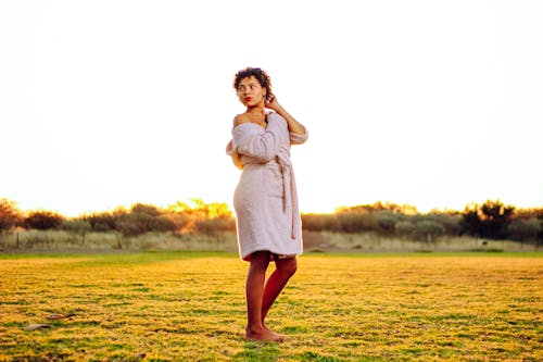 Side view of pensive young barefooted ethnic female in bathrobe touching curly hair and looking over shoulder while standing on grassy meadow near green trees on sunny day