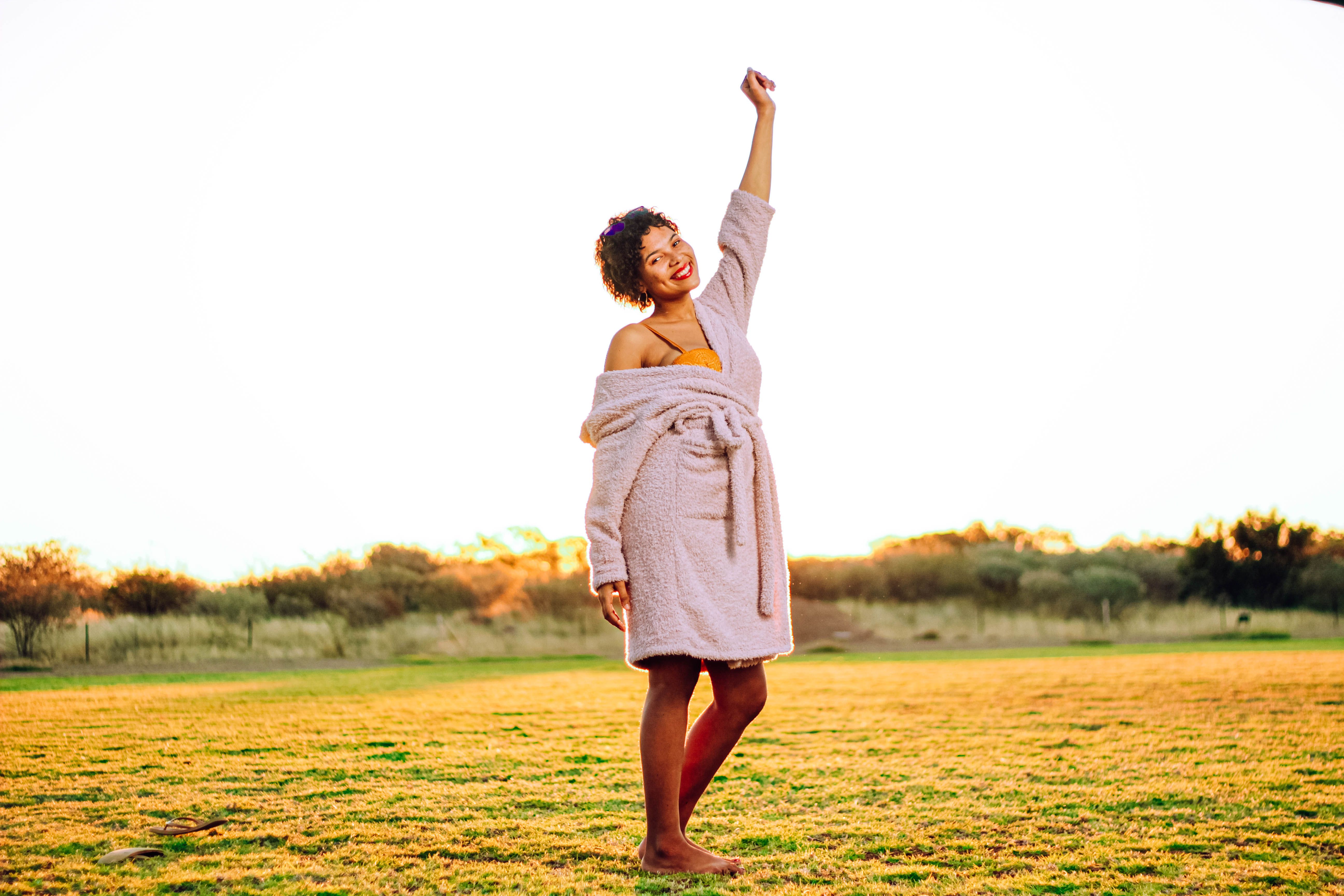 A woman standing in a sunny meadow with one arm raised, smiling