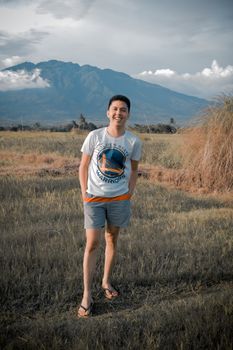 A young man smiles in a field with a mountain backdrop, dressed casually in a t-shirt and shorts.