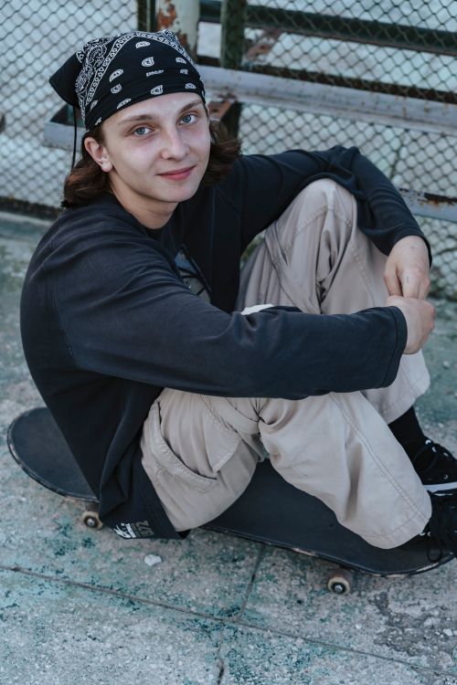 Free A teenage skateboarder in a bandana sits on a skateboard, smiling on a pavement Stock Photo
