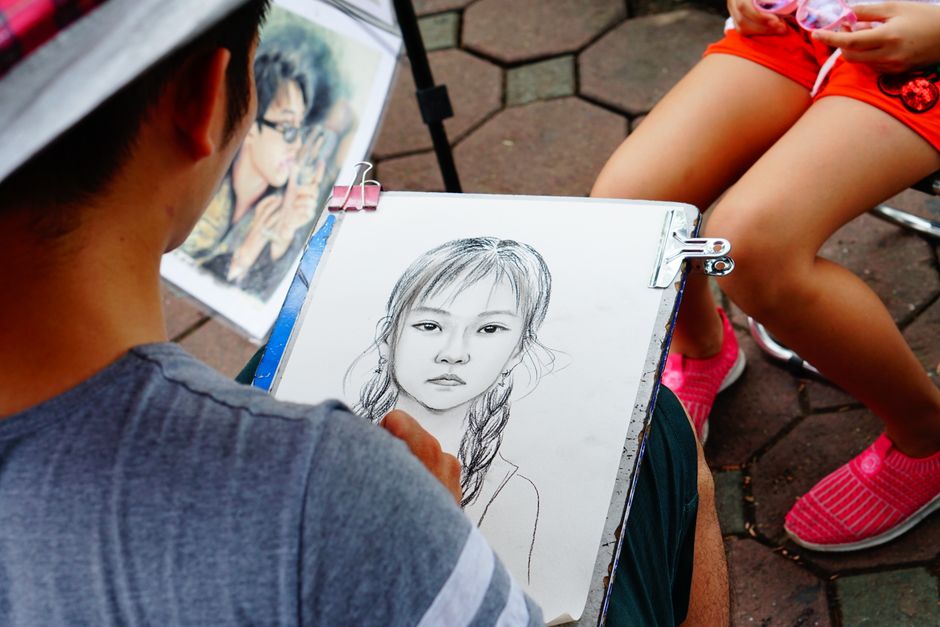 A street artist creates a pencil portrait of a young girl in an outdoor setting.