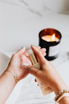 Close-up of a woman applying skincare lotion with a burning candle in the background, creating a cozy ambiance.