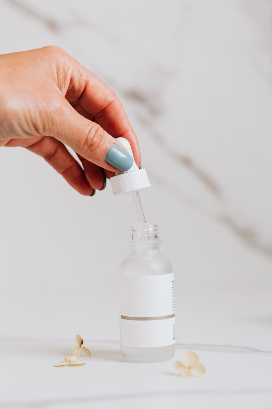 A hand holding a pipette above a glass dropper bottle, applying a skincare serum in soft natural light