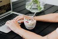 一个人坐在笔记本电脑前处理多项任务，木桌上放着一份营养酸奶冻糕，里面有新鲜水果和格兰诺拉麦片