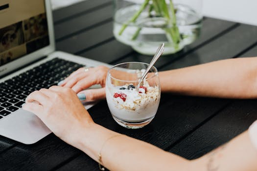 Person multitasking at a laptop with a nutritious yogurt parfait, featuring fresh fruits and granola, on a wooden table.