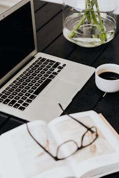 整洁的工作空间里，深色桌子上摆放着笔记本电脑、咖啡杯、打开的书和一副眼镜