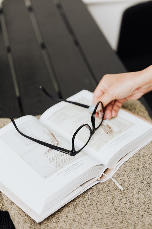 Free A hand holds glasses over an open book on a table, capturing a moment of study or reading. Stock Photo