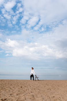 A lone man dressed in white and black stands on a tranquil sandy beach under a blue sky.