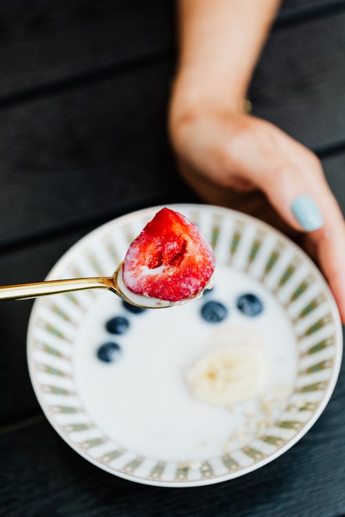 Free A hand holding a gold spoon with a strawberry, in a bowl with milk, blueberries, and banana. Stock Photo