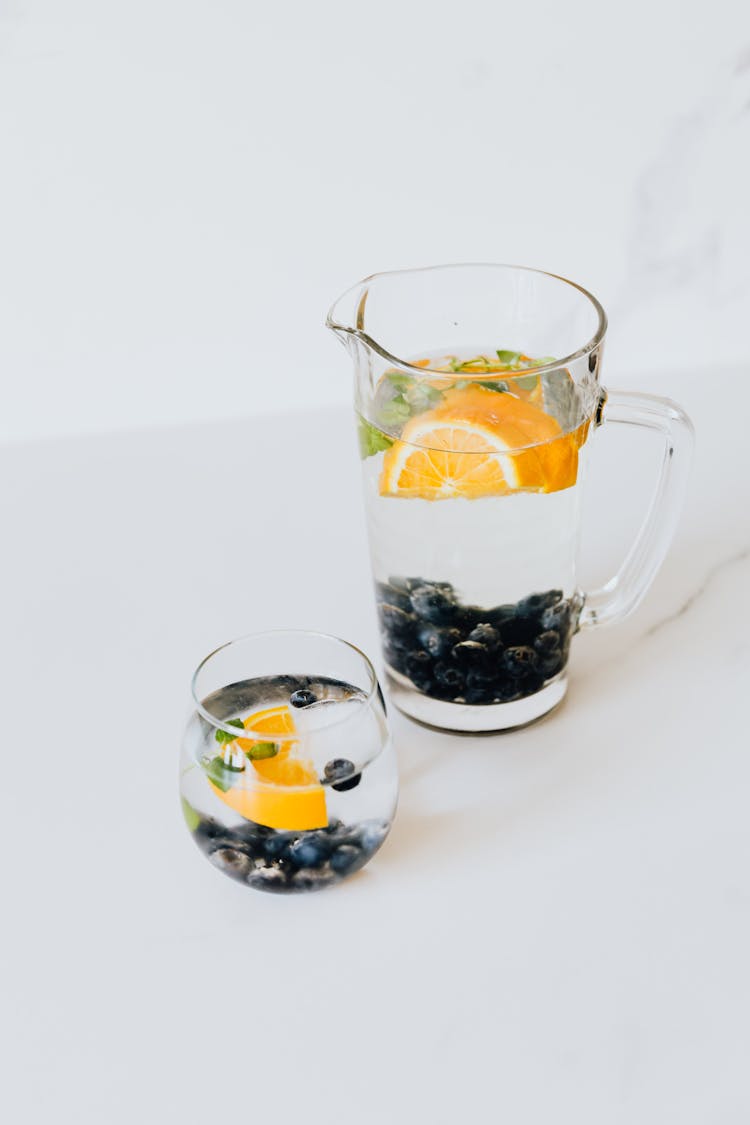 A Clear Glass And A Pitcher With Slices Of Orange Fruit And Blueberries In Water