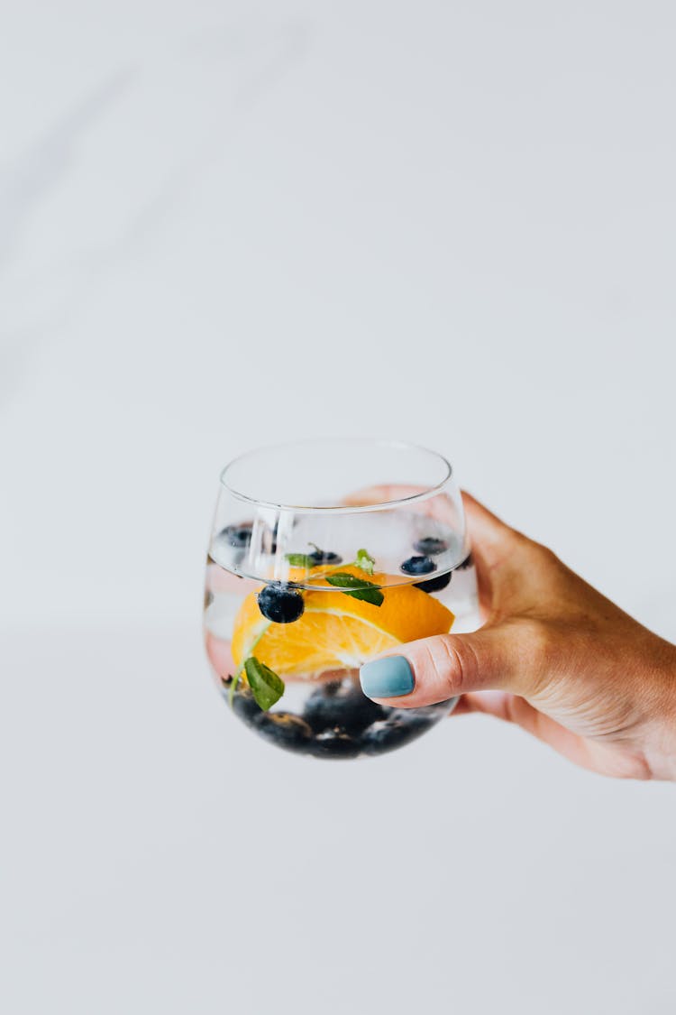 A Person Holding A Glass Of Water With Blueberries And A Slice Of Orange Fruit