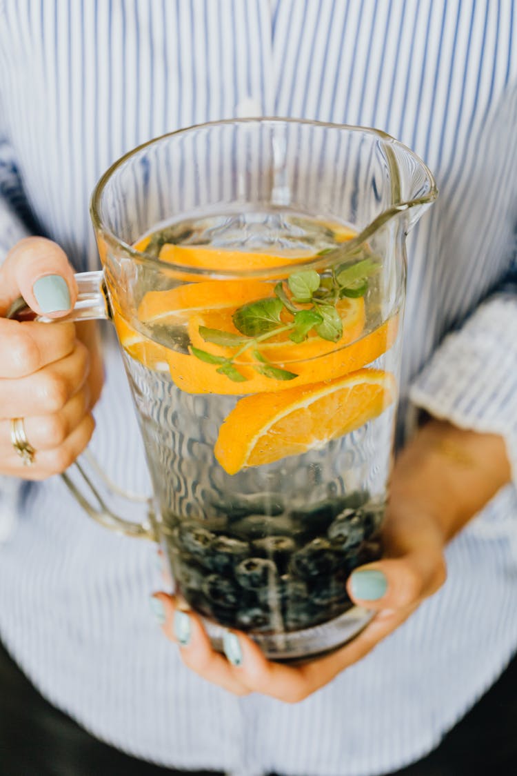 A Person Holding A Pitcher Of Water With Slices Of Orange, Blueberries And Mint Leaves