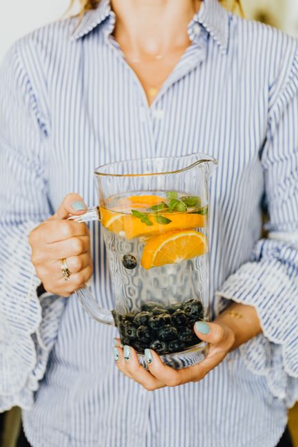 A glass pitcher of infused water filled with lemon slices, cucumber rounds, and mint leaves, sitting on a wood countertop next to two filled glasses with ice.