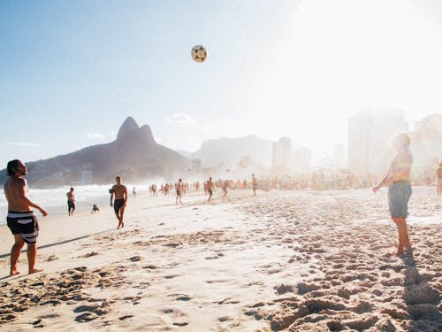 Group of people playing volleyball on crowded shore near calm sea against mountainous terrain under sunlight in summer time in resort