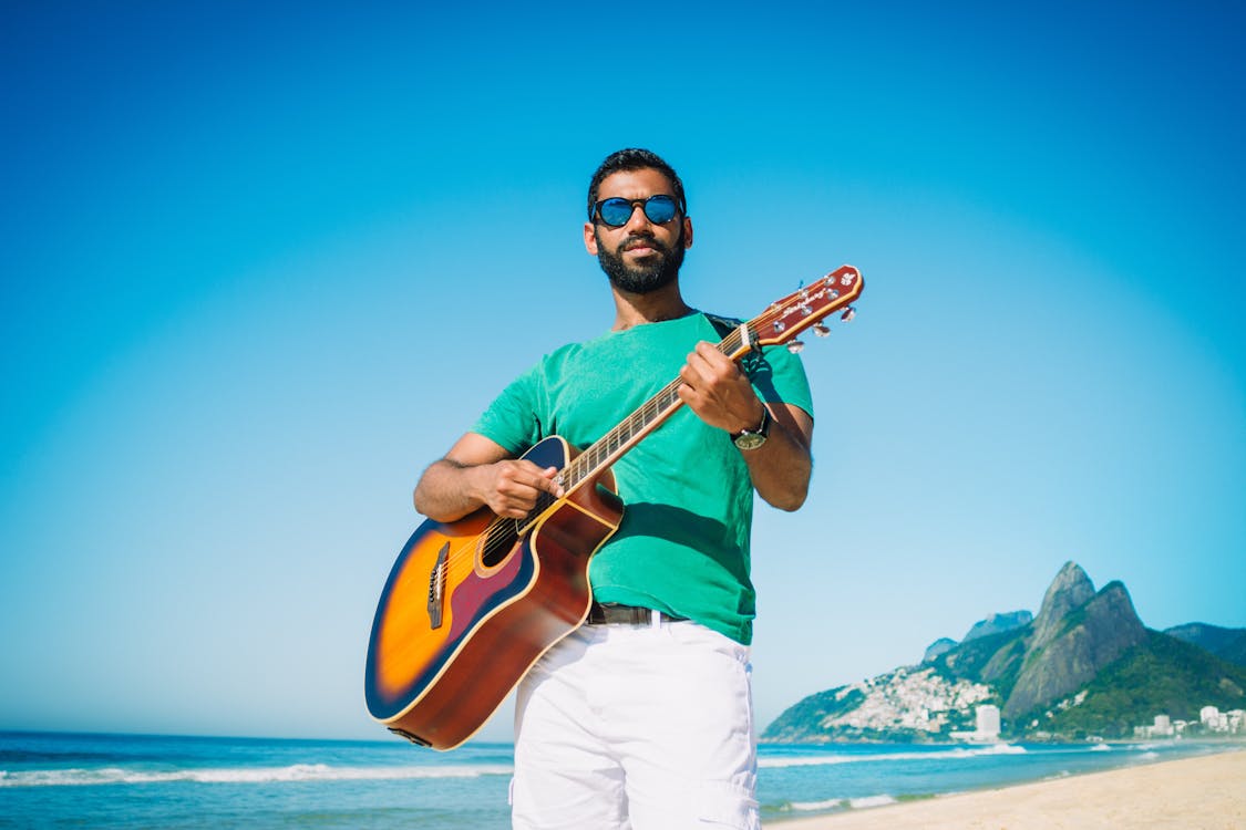 Low angle of content male in sunglasses playing acoustic guitar while standing on sandy beach near turquoise sea and looking at camera