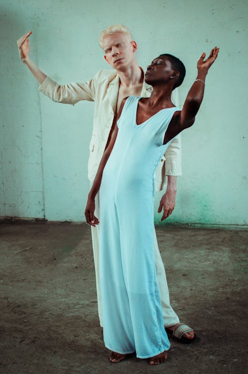 Full body of multiethnic couple of dancers in white outfit standing with eyes closed and arms raised near white wall