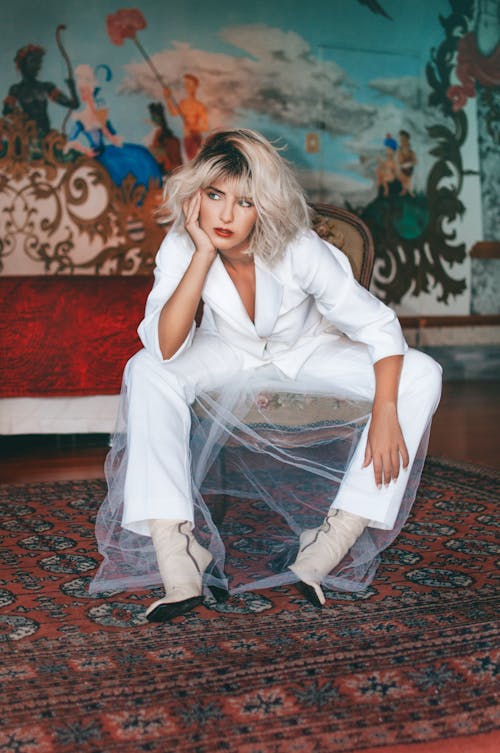 Beautiful female with blond hair wearing white costume and high heel beige boots sitting on ornamental chair while leaning on hand and looking away