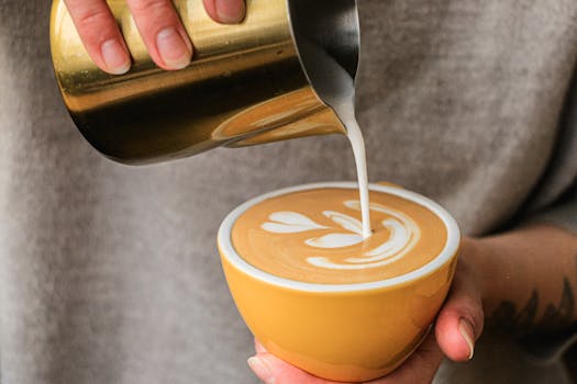 Close-up of a hand pouring steamed milk into a latte with art in a ceramic cup.