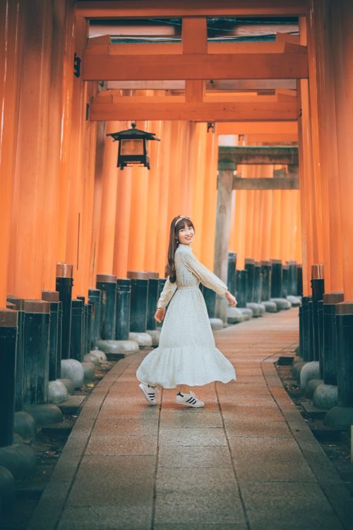 Free A woman in a white dress smiling while walking through the iconic orange torii gates of Fushimi Inari Shrine. Stock Photo