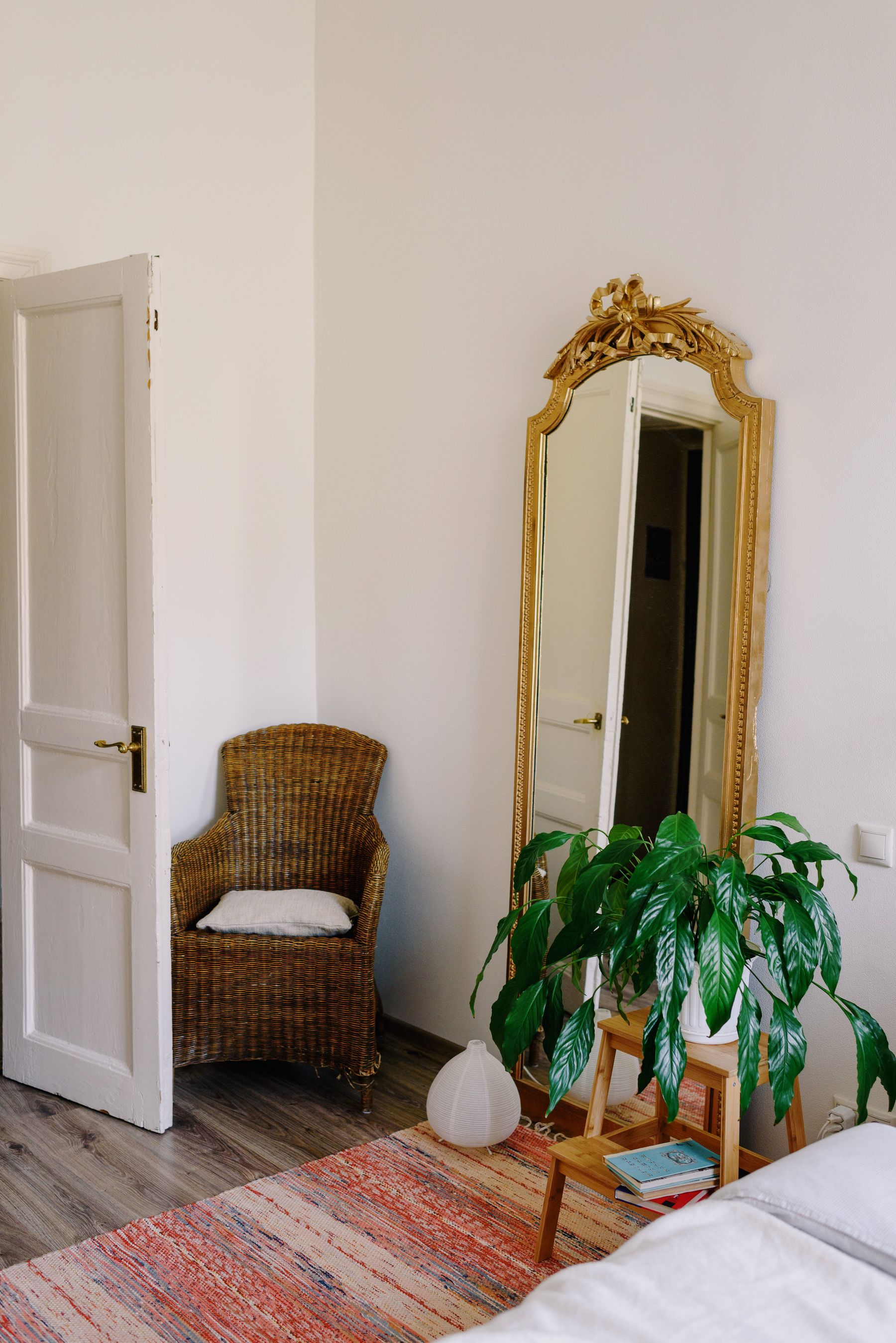 Bedroom corner with a wicker chair, tall mirror, ornamental rug and potted plant beside the bed