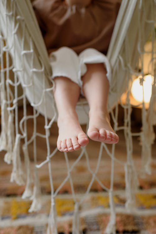 Crop unrecognizable barefoot kid sitting in hammock with fringe above floor at home in daylight