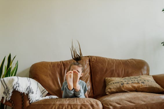 Anonymous little barefoot kid with flying hair having fun on comfortable leather couch in house