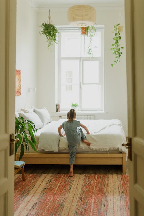 Back view of unrecognizable little barefoot kid in pajamas climbing onto soft bed in light house
