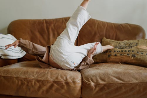 Unrecognizable little child with raised legs jumping on comfortable couch at home in daylight