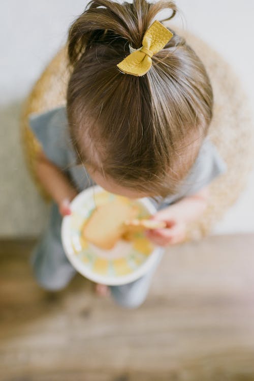 From above of unrecognizable little girl eating crunchy biscuit while sitting on stool at home