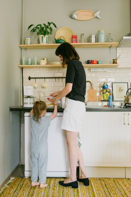 Full body of anonymous young woman in casual clothes preparing breakfast for little daughter in pajama while standing together in kitchen