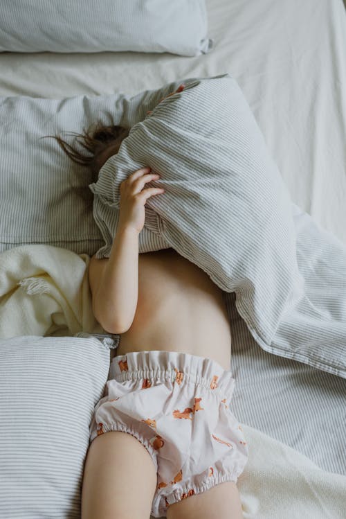 Free From above of unrecognizable adorable little child in panties covering face with blanket while lying on comfortable bed in morning Stock Photo