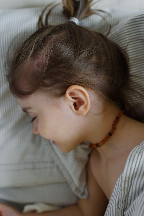 Free From above side view of adorable little girl with beaded necklace lying on soft bed with closed eyes and smiling Stock Photo