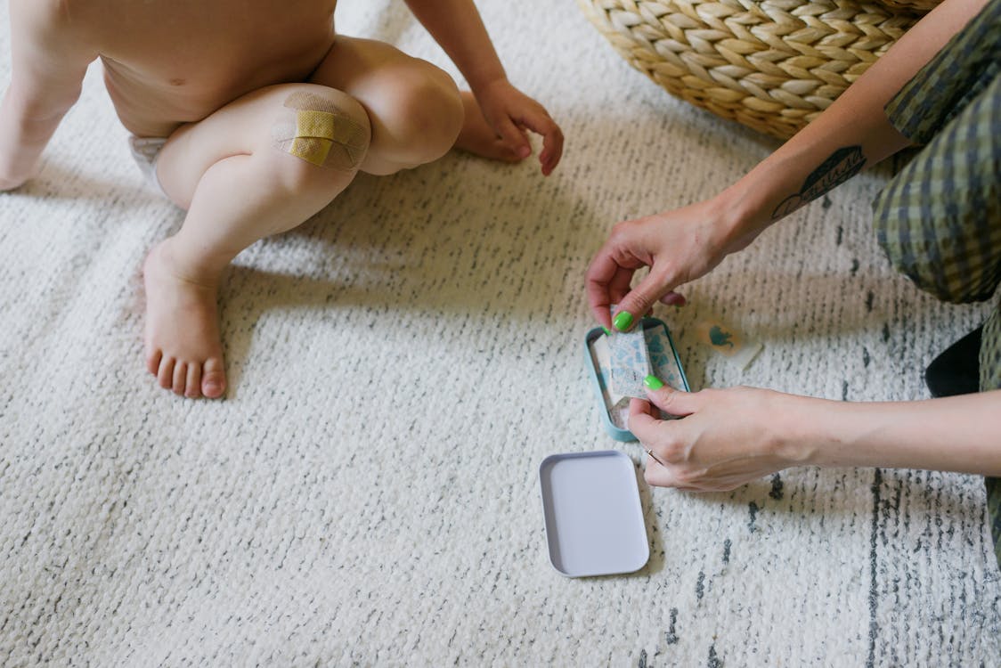 From above of crop anonymous barefooted little child with wound on knee sitting on carpet and choosing adhesive plaster with unrecognizable tattooed mother