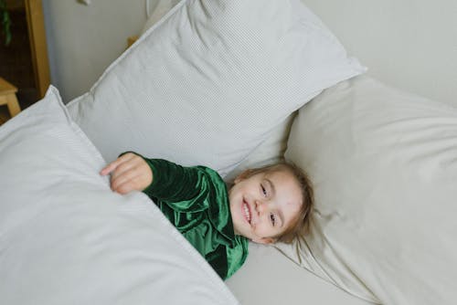 Free From above of adorable happy little girl in green dress smiling and looking at camera while lying on bed under soft blanket Stock Photo
