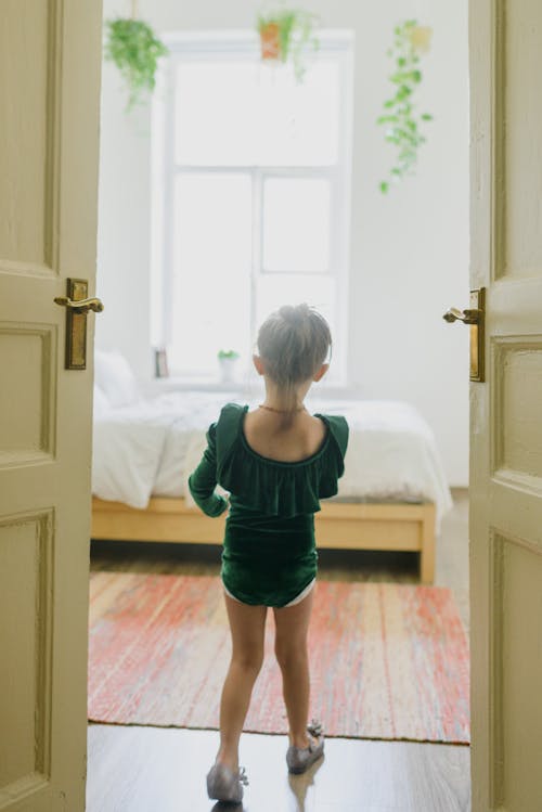 Back view of anonymous cute child in stylish green bodysuit walking in cozy bedroom on sunny day