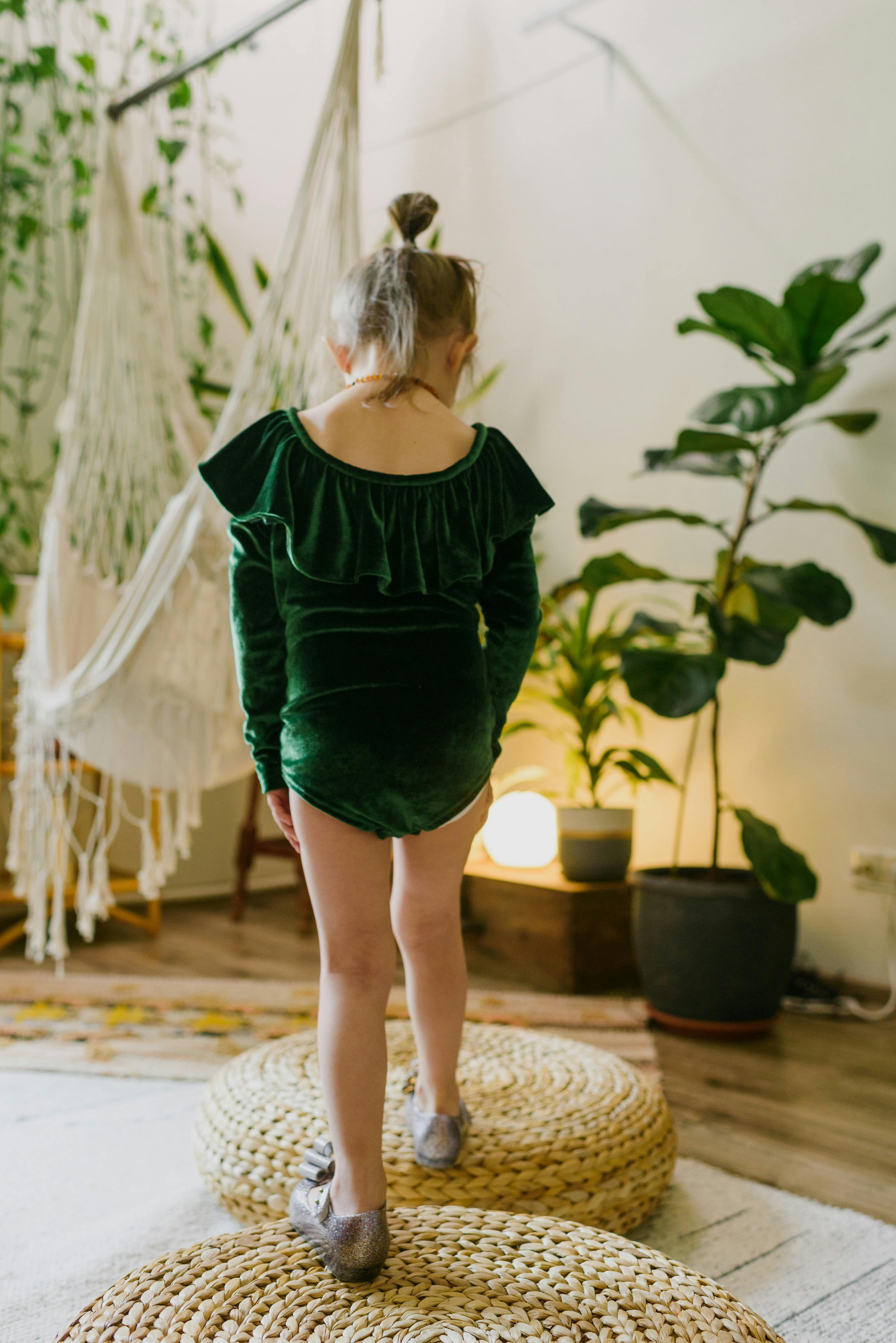 Back view of unrecognizable stylish little girl in green costume standing on traditional rattan weaved poufs placed in cozy apartment in sunlight