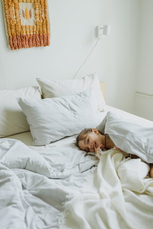 Free Smiling little girl hiding under blanket while playing on bed Stock Photo