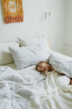 From above of happy curious little kid lying under blanket on bed and smiling in sunny morning